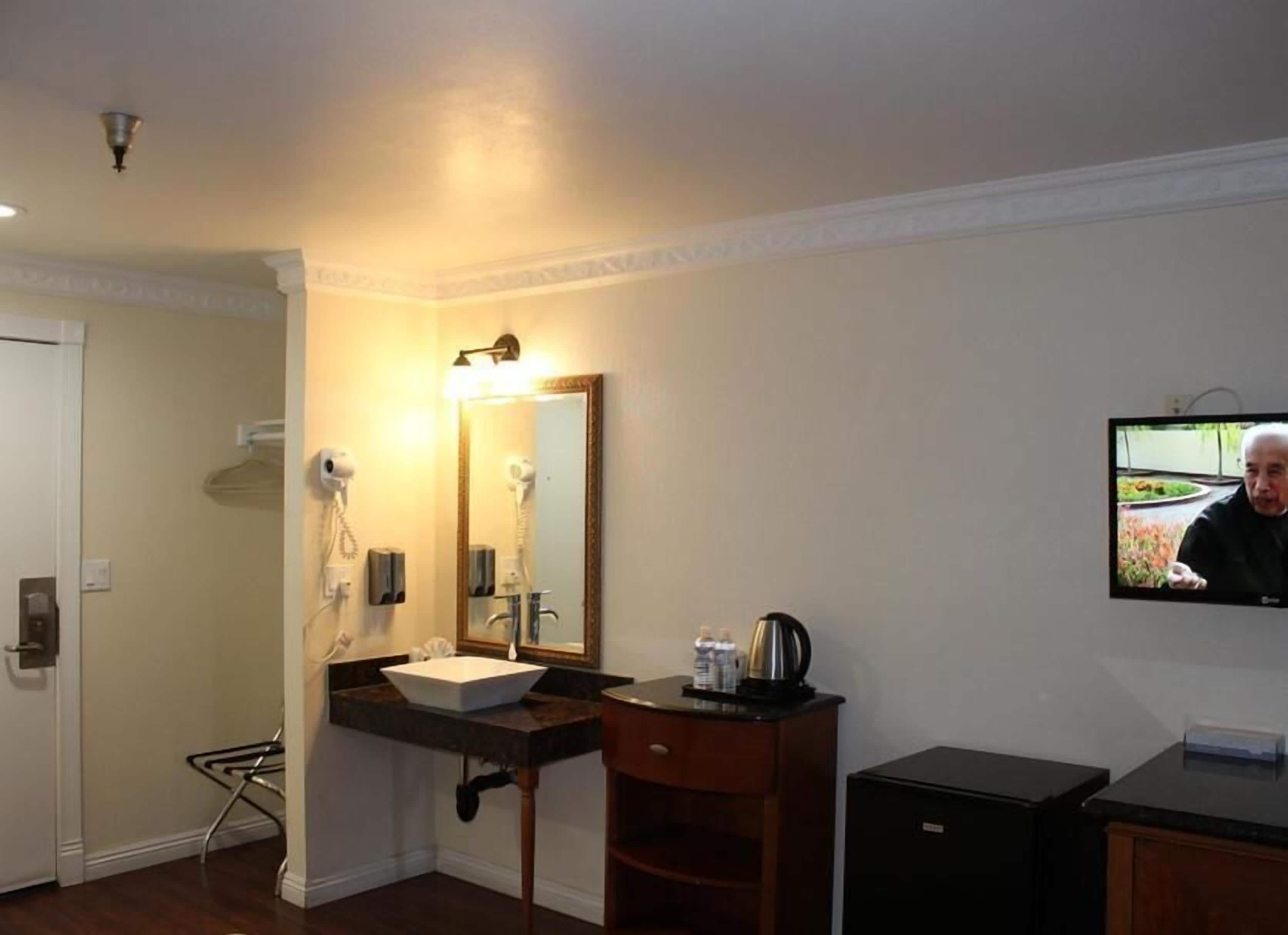 A hotel room with a bathroom featuring a sink, mirror, and hairdryer. There is a television mounted on the wall and a small cabinet.