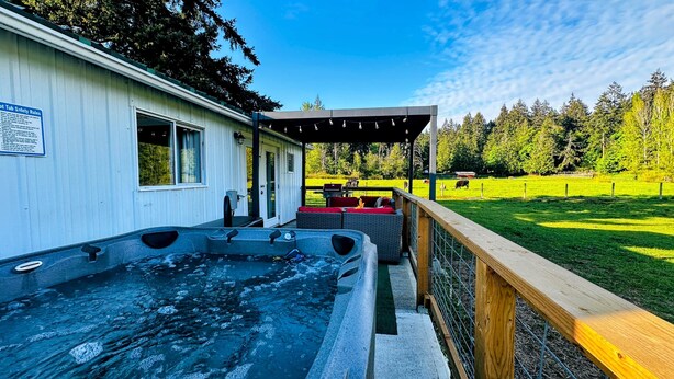 A hot tub outside a building with a deck, a covered seating area, and a fenced yard with trees.