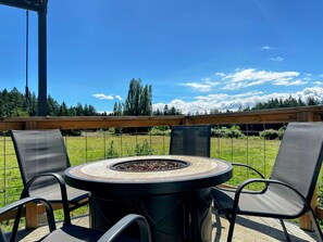 An outdoor patio with a round table and chairs, a fire pit, and a view of a grassy field and trees under a clear sky. - The Cottage on the Farm (Ladysmith)