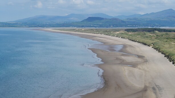 A coastal landscape with a wide sandy beach, gentle waves, and distant mountains under a clear sky.