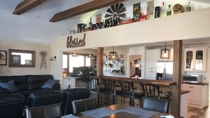 Un comedor con una mesa larga preparada para cuatro personas, una zona de bar con taburetes y una cocina a la vista al fondo.