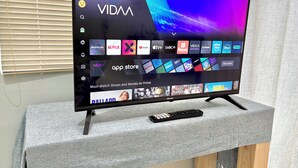 A flat-screen TV on a stand displaying a VIDAA interface with various streaming apps and a remote control on a grey table. - One Bedroom Bachelor A (Cape Town)