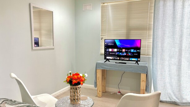 A room with a television on a stand, a small table with a vase of flowers, and two white chairs. - One Bedroom Bachelor A (Cape Town)