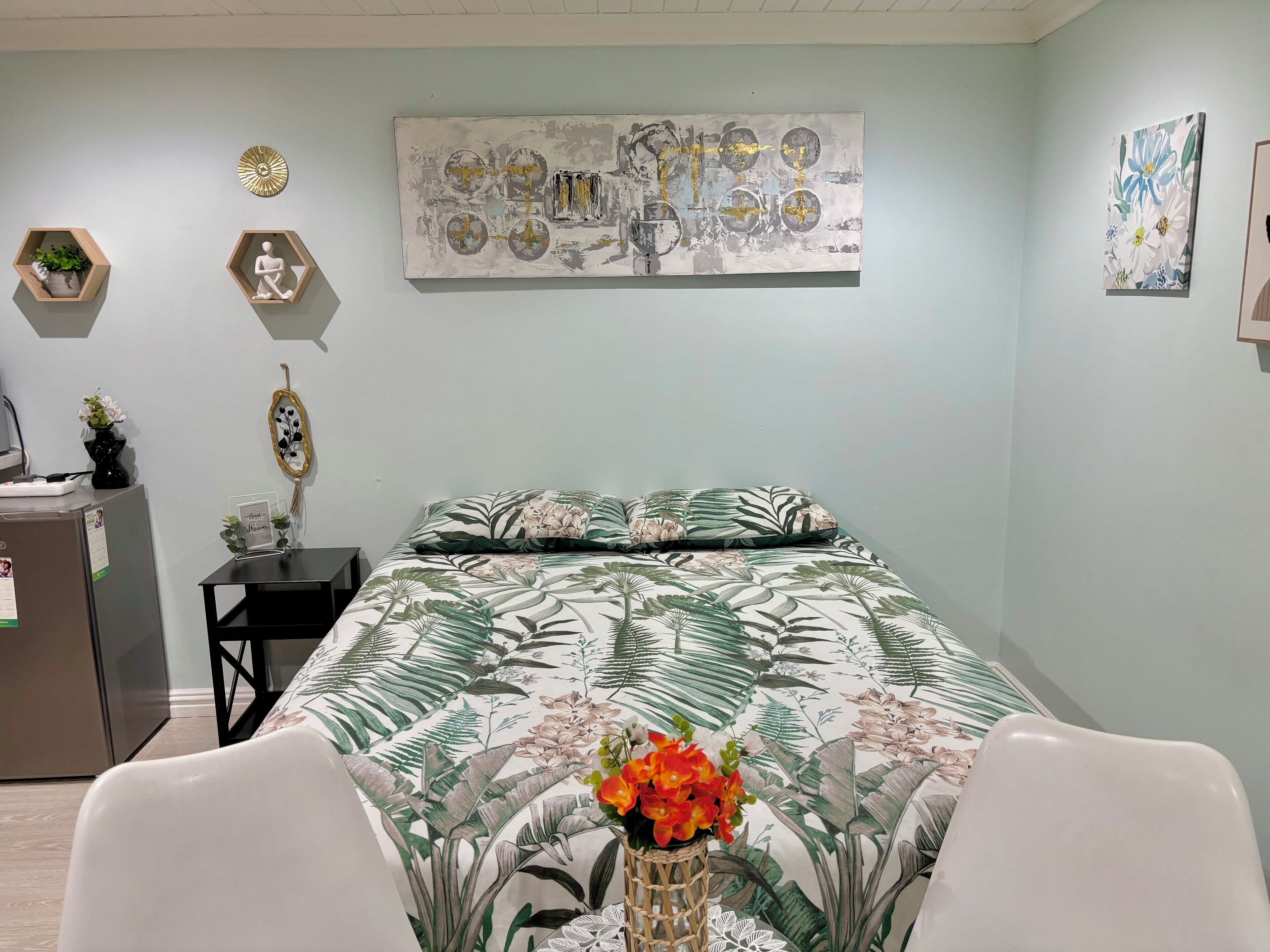 Un dormitorio con cama, escritorio y refrigerador. Hay dos repisas hexagonales en la pared, una con una planta y la otra con un objeto decorativo.