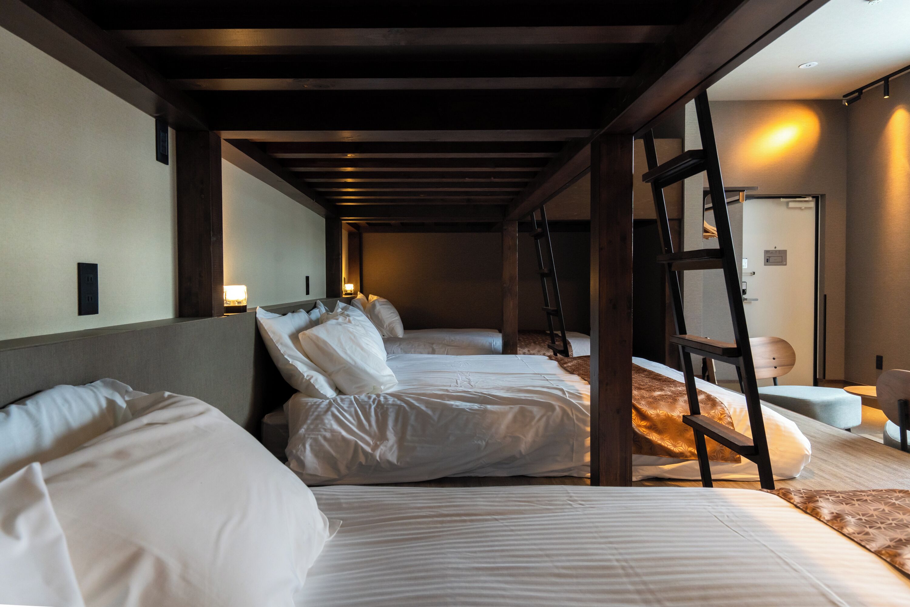 A hotel room with three beds, wooden beams, a ladder, and a small table with a lamp.