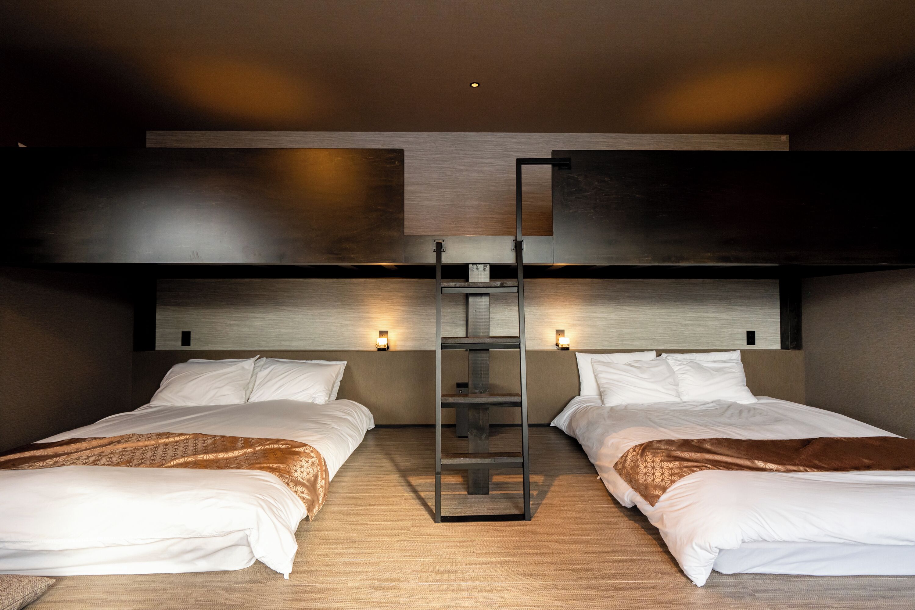 A modern hotel room with two beds, a central ladder, and wall-mounted lights.