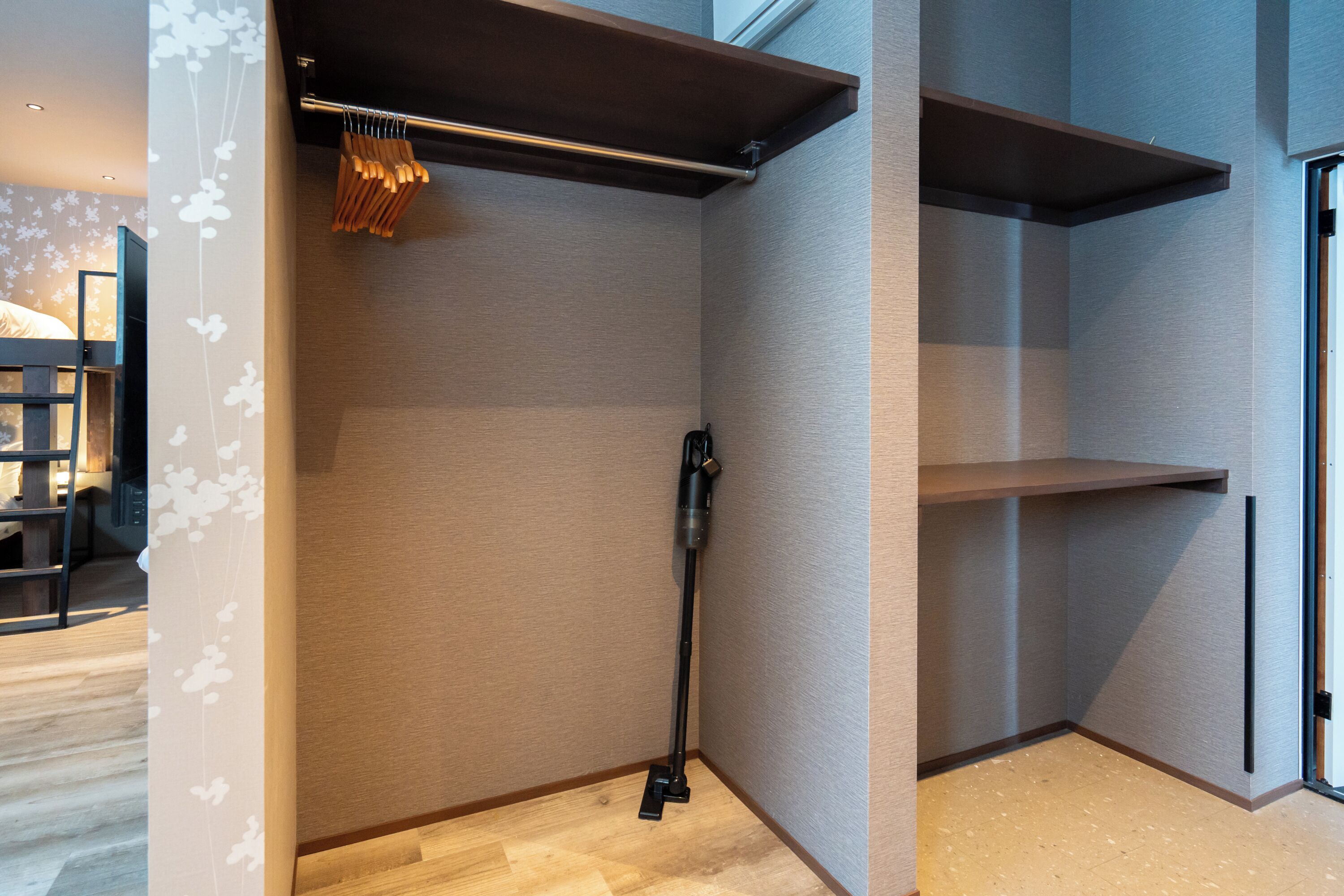 A corner of a room with a wooden floor, a black vacuum cleaner, and a wooden shelf unit.