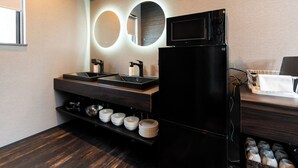 Una cocina moderna con un refrigerador negro, un lavamanos y un microondas. Hay estantes con cuencos y platos, y un espejo con luces circulares.
