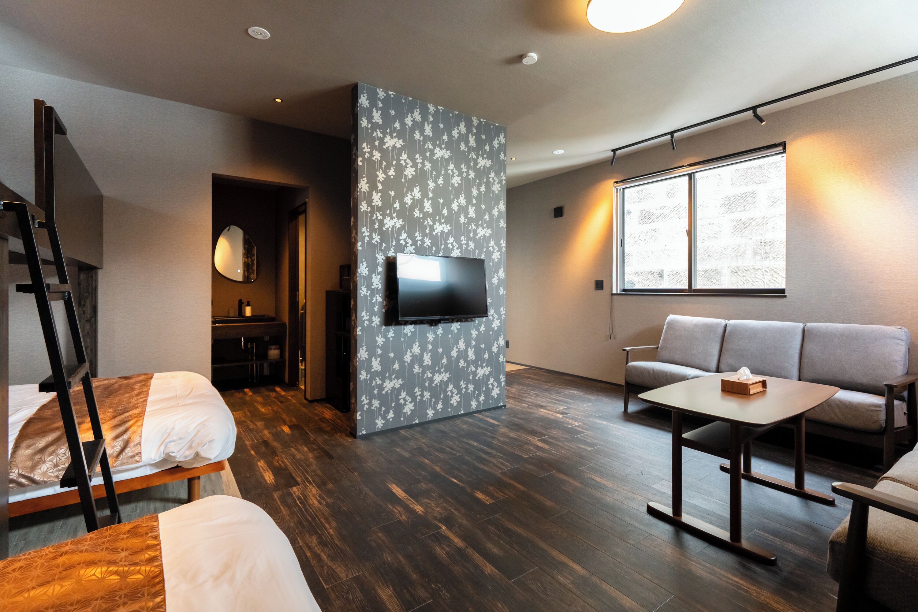 Ein modernes Hotelzimmer mit Holzboden, einer Couch, einem an einer blumengemusterten Wand montierten Fernseher und einem Fenster mit Tageslicht.