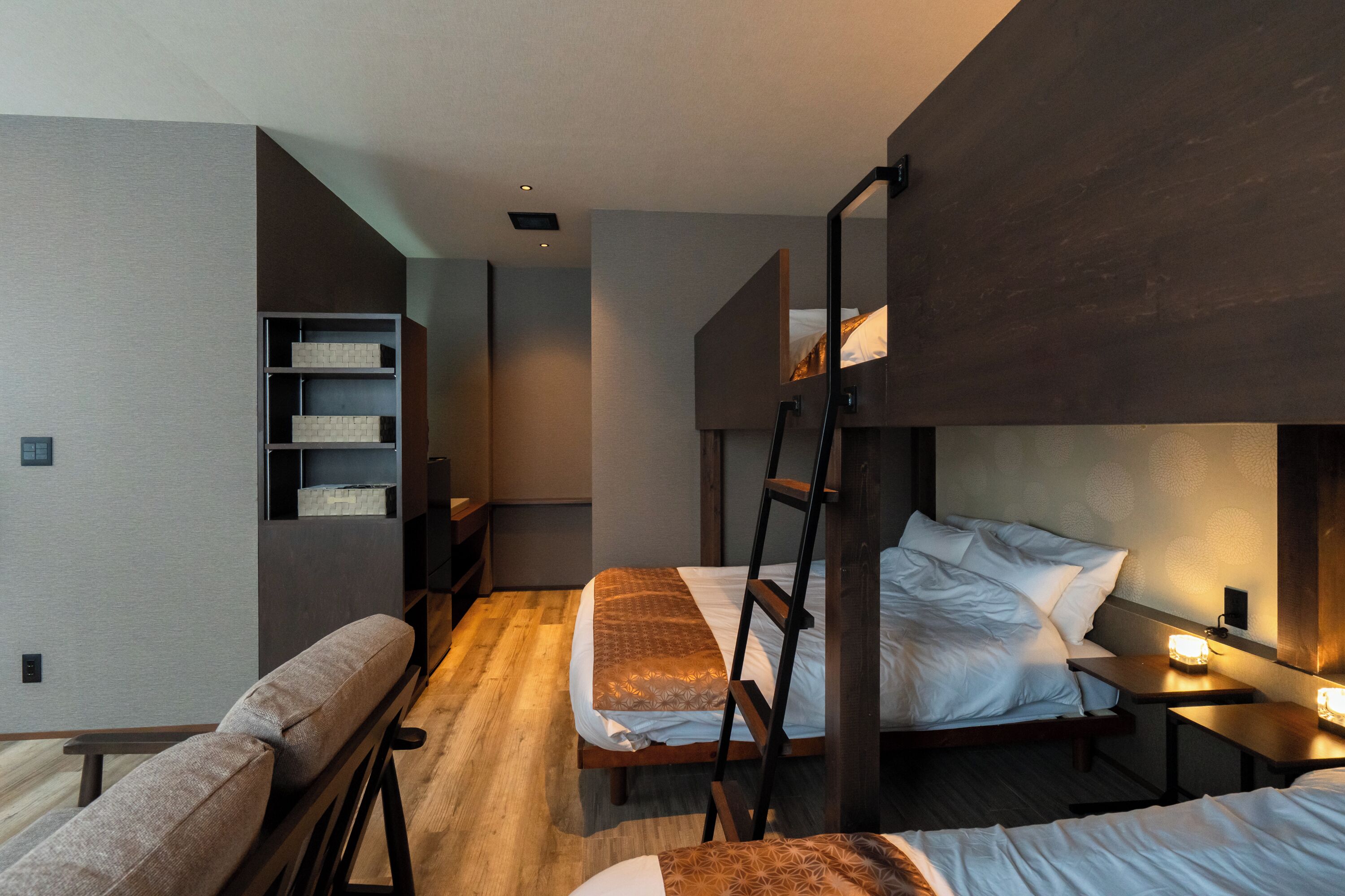 A modern hotel room with a bunk bed, a sofa, a ladder, and a bedside table with a lamp.