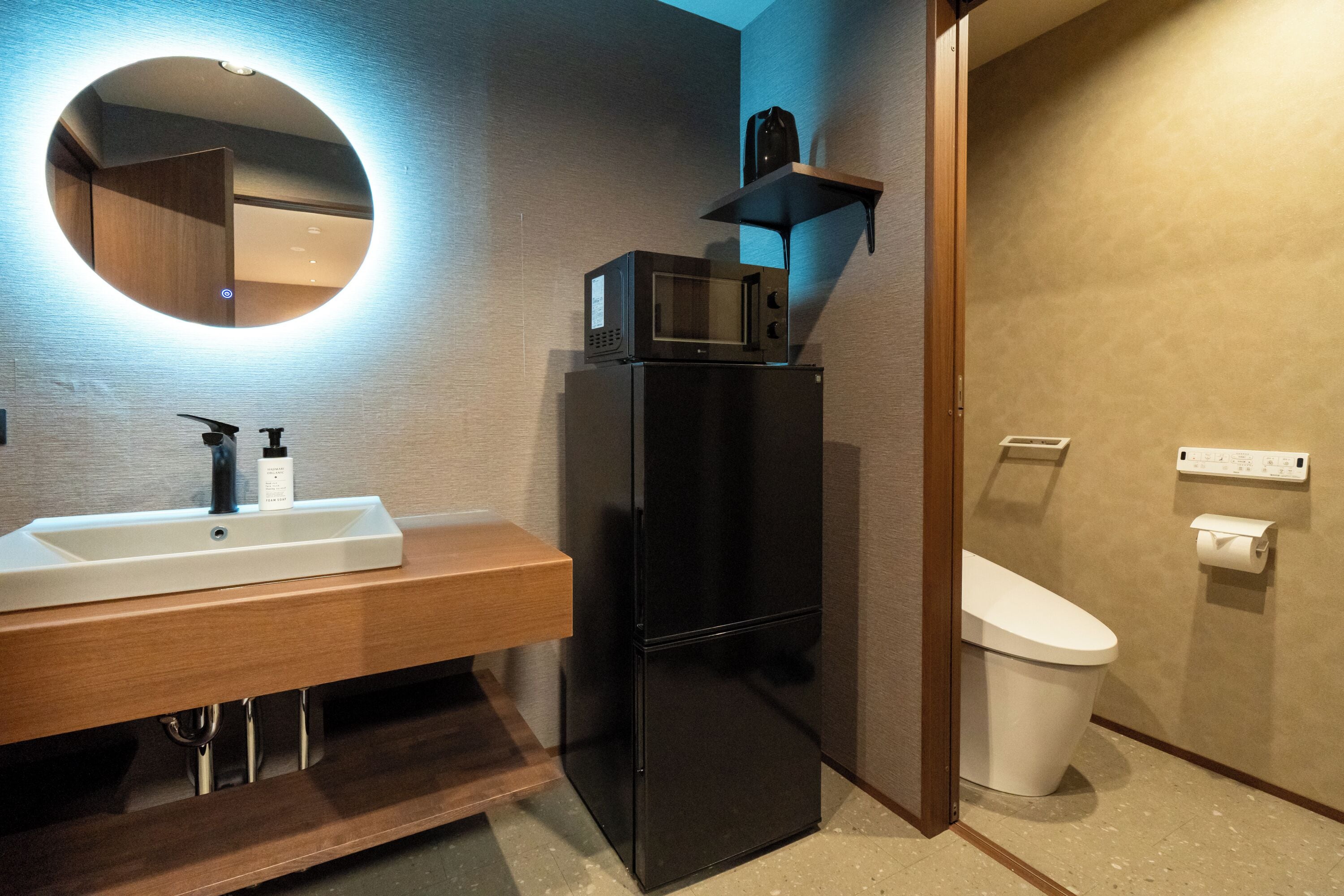A modern bathroom with a black refrigerator, a sink with a soap dispenser, a mirror with a circular light, and a toilet with a paper holder.