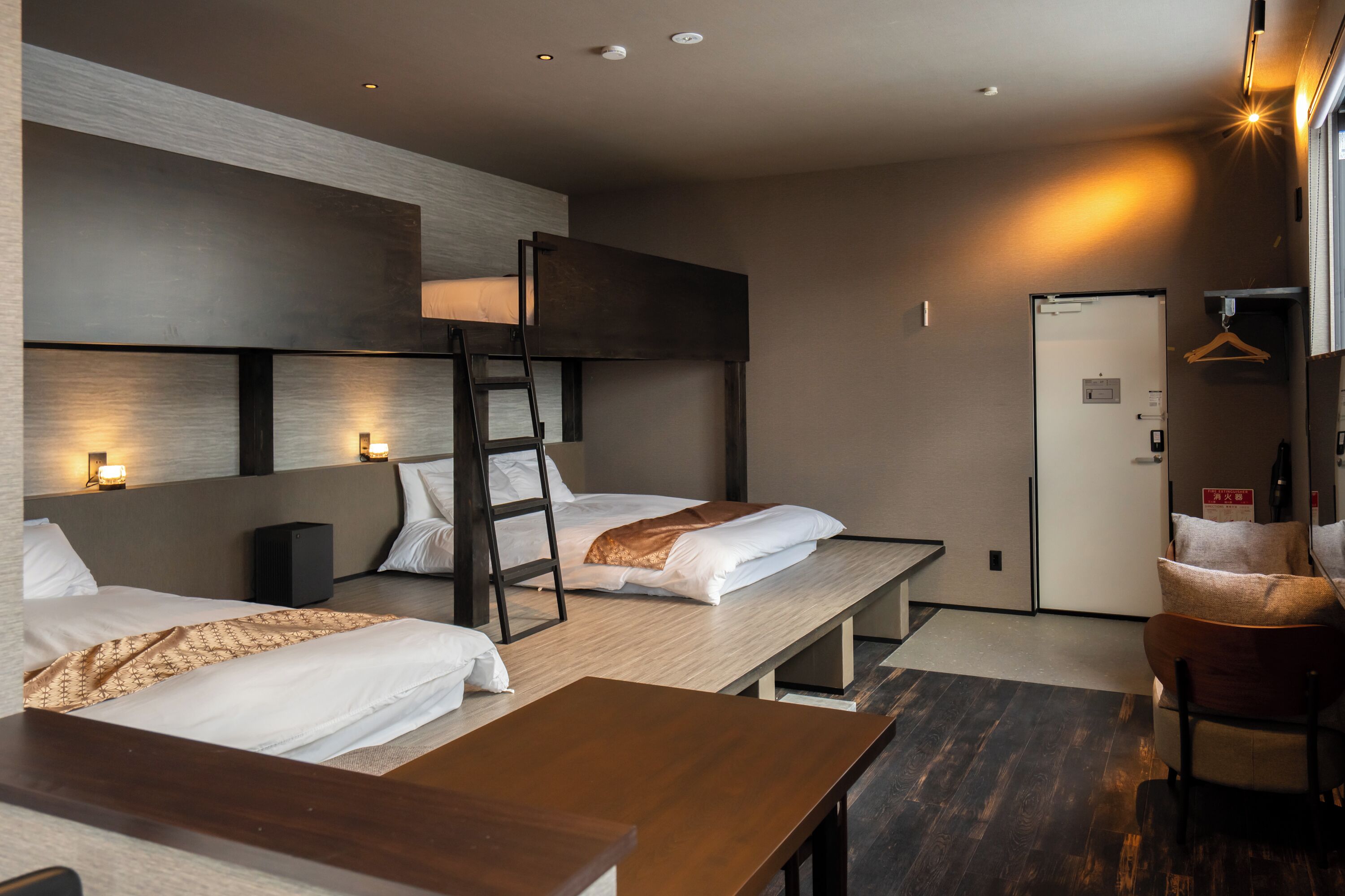 A hotel room with bunk beds, a desk, and a chair. There is a door, a coat hanger, and a small table.