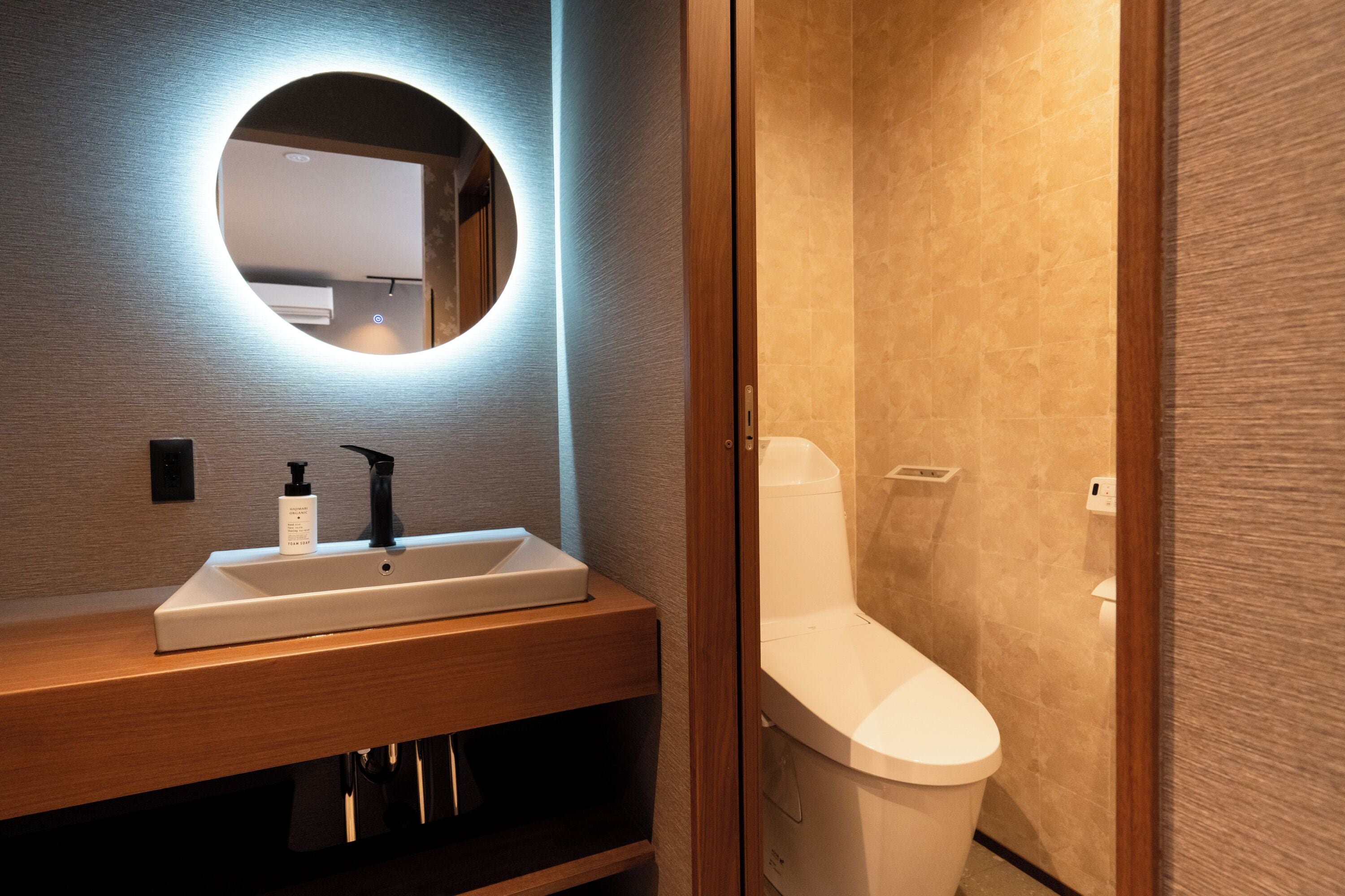 A modern bathroom with a wooden vanity, a white sink, a black faucet, a round mirror with a light, and a white toilet.
