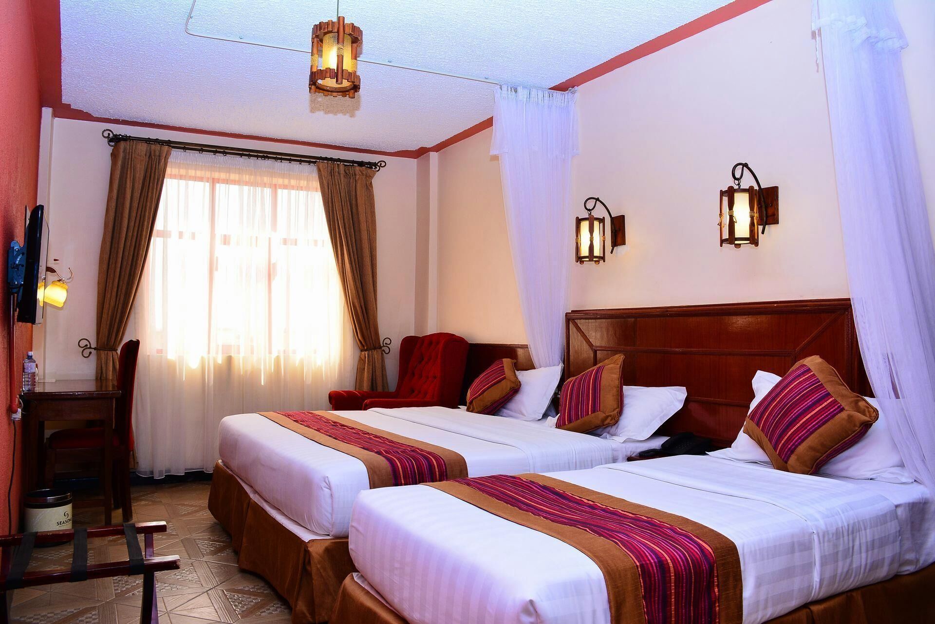 Habitación de hotel con dos camas, cabecero de madera, ventana con cortinas y dos lámparas de pared.