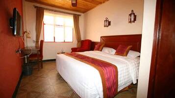 Une chambre d’hôtel avec un plafond en bois, un lit avec une literie blanche et un élément décoratif rouge, une chaise, un bureau avec une lampe et une fenêtre avec des rideaux.