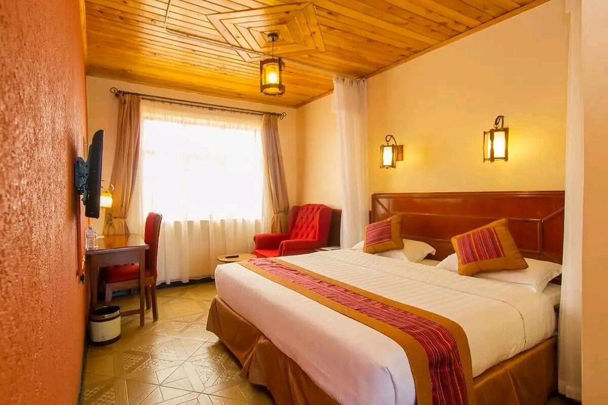 Une chambre d’hôtel avec un plafond en bois, un grand lit recouvert d’une courtepointe rouge et blanche, deux lampes fixées au mur, un bureau avec une chaise et une fenêtre avec des rideaux.