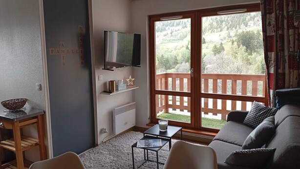 A living room with a TV, a wooden table, a sofa, and a balcony with a wooden railing. - Apartment at the foot of the slopes, near outdoor swimming pool (summer) with garage. (Saint-François-Longchamp)