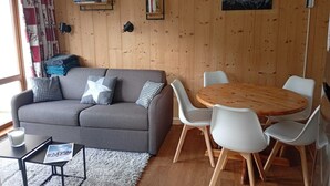 A cozy living room with a grey sofa, white chairs, a wooden dining table, and a bookshelf against a wooden wall. - Apartment at the foot of the slopes, near outdoor swimming pool (summer) with garage. (Saint-François-Longchamp)