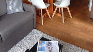 A cozy living room with a wooden wall, a round wooden table with four white chairs, a grey sofa with a star-patterned pillow, and a door in the background. - Apartment at the foot of the slopes, near outdoor swimming pool (summer) with garage. (Saint-François-Longchamp)