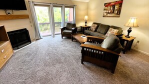 A living room with a fireplace, a television mounted on the wall, a sofa, a chair, and a coffee table. There is a painting on the wall and a window with curtains. - Nordic Village | Arlberg Two-Level Condo w/ Jetted Tub (Jackson)