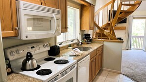 A compact kitchen with a microwave, stove, and dishwasher. It features wooden cabinets and a small window with blinds. - Nordic Village | Arlberg Two-Level Condo w/ Jetted Tub (Jackson)