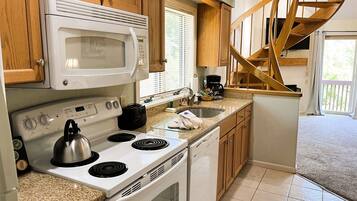 A compact kitchen with a microwave, stove, and dishwasher. It features wooden cabinets and a small window with blinds.