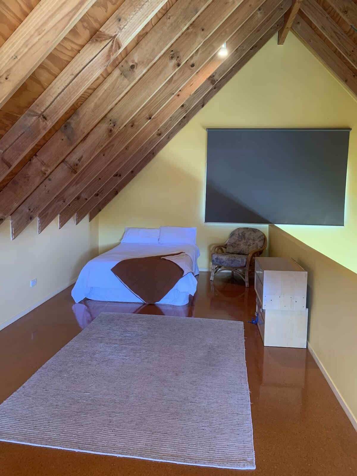 A bedroom with a sloped ceiling, a bed, a chair, and a small white cabinet.