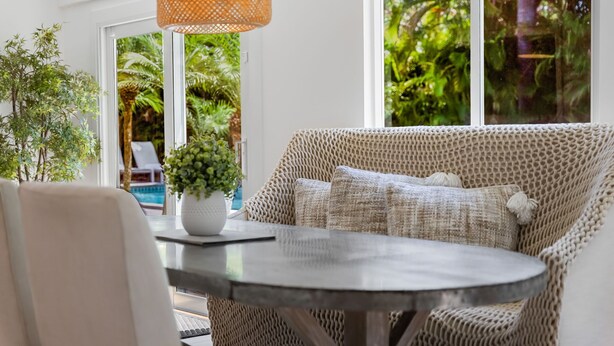 A cozy seating area with a woven sofa and a round table, a potted plant, and a view of lush greenery outside.