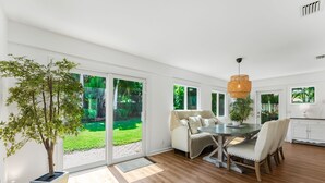 A spacious room with a large sliding glass door leading to a garden, a dining area with a round table and chairs, and a kitchen area with white cabinets.