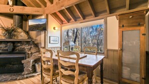 Un coin repas rustique avec une cheminée en pierre, des chaises en bois et une vue sur les arbres à travers de grandes fenêtres.