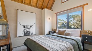 Une chambre à coucher avec un grand lit, une table de chevet, une lampe et une fenêtre donnant sur des maisons et des arbres.
