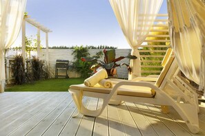Un espacio de estar al aire libre techado con una silla de lounge y una mesa, una parrilla y una pérgola con cortinas blancas.