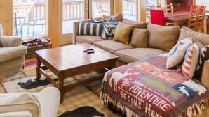 Un salon spacieux avec un grand canapé, une table basse et une cheminée. De nombreuses fenêtres laissent entrer la lumière naturelle et offrent une vue sur l’extérieur.