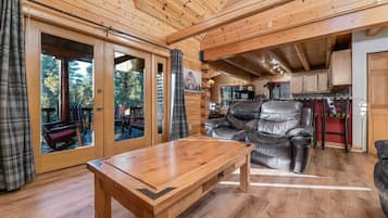 A wooden cabin interior with a large sliding glass door leading to an outdoor deck, a leather sofa, and a wooden table.