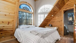 A bedroom with a sloped ceiling, wooden walls, a large bed with striped bedding, a window with a decorative metal frame, and a small bedside table with books. - Indulge in the Perfect Family Adventure (Sugarloaf)