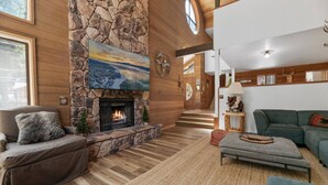 A cozy living room with a stone fireplace, a large painting, and a staircase leading to another level. - The Circle Moon - 1 mi to Bear Mtn/ Game Room+Spa! (Big Bear Lake)
