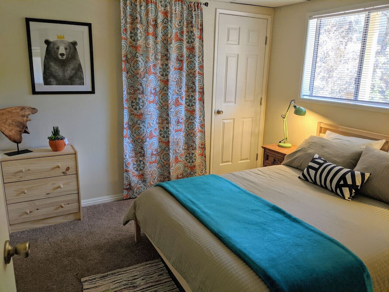 A bedroom with a bed, a nightstand, a dresser, a window with blinds, a door, and a wall with a framed picture.