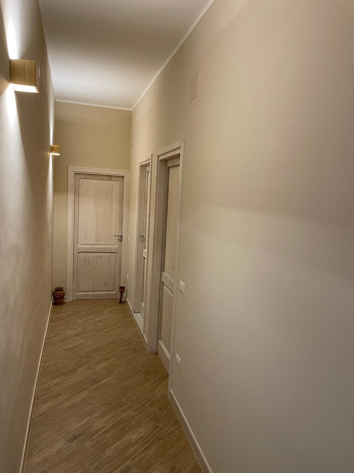 Un pasillo estrecho con suelo de madera y paredes blancas. Se observan tres puertas a la derecha y un objeto rojo pequeño en el suelo cerca de la pared izquierda.