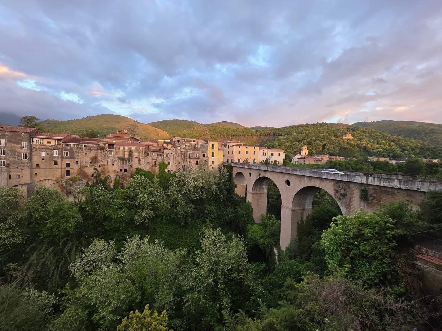 Un puente de piedra histórico con múltiples arcos que cruza un río, rodeado de abundante vegetación y con colinas al fondo.