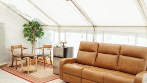 A tented room with a brown leather sofa, two wooden chairs, a small table, and a refrigerator.
