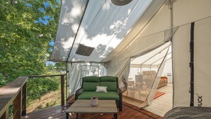 A tent with a green sofa and a table, a wooden deck, and a view of trees.