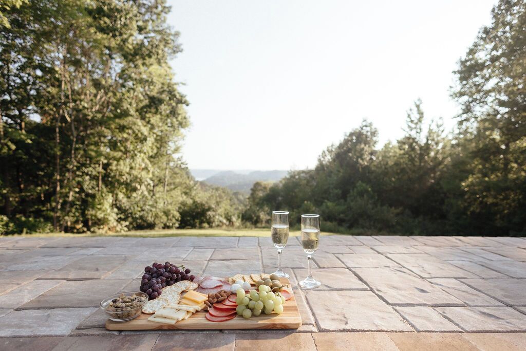 A picnic setup with a variety of cheeses, fruits, and nuts on a wooden board, accompanied by two glasses of white wine, set on a stone table outdoors with a forested background.