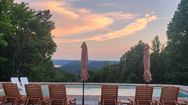 A poolside area with wooden chairs and umbrellas, overlooking a scenic landscape with trees and hills.