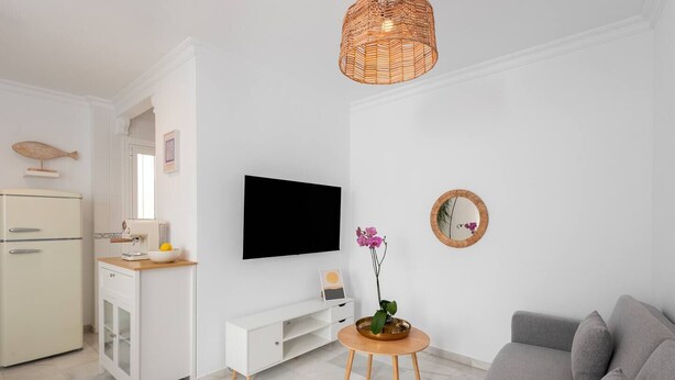 A modern living room with a grey sofa, a television, a small wooden side table with a vase, and a kitchen area with a fridge and a microwave. - Centric but quiet. Terrace with views to the sea (Nerja)