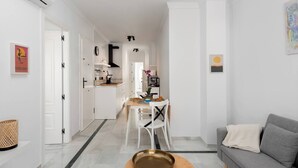 Una cocina moderna con armarios blancos, un comedor con mesa y sillas, y un sofá gris en un luminoso salón con paredes blancas.