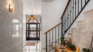 Un pasillo con piso de mármol, un banco de madera con revistas, una escalera negra y una puerta negra con un panel de vidrio.
