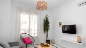 A modern living room with a grey sofa, a wicker chair with a pink cushion, a round wooden table, a potted plant, a TV on the wall, and a white air conditioning unit. - Main street free parking 5 minutes away (Nerja)