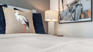 Un lit avec une couette blanche, un oreiller orné d’un motif d’oiseau, une lampe et un tableau accroché au mur.