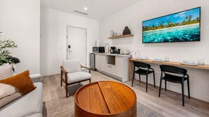 Un salon moderne avec une table basse ronde en bois, un canapé et deux fauteuils. On aperçoit à l’arrière-plan un coin cuisine équipé d’un four à micro-ondes, d’un grille-pain et d’une machine à café.