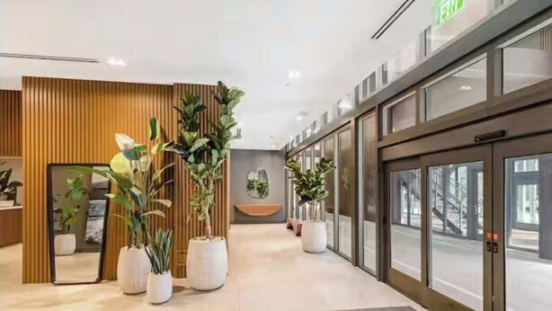 Un hall d’hôtel moderne avec un comptoir d’accueil en bois, des plantes en pot et un ascenseur en verre.